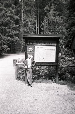 Vintage black-and-white photo of a man posing beside a wooden informational board in a forested area. The sign reads *"Boubin...