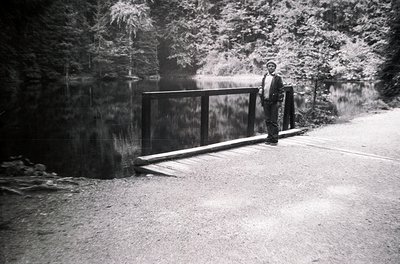 Black-and-white mid-century roadside scene featuring a man in a suit and hat standing beside a modernist metal bridge over a ...