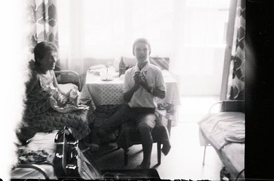 Vintage black-and-white indoor scene featuring two seated individuals in a modest room. One holds a small child, while the ot...