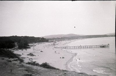 Vintage black-and-white seaside scene featuring a long pier extending into calm waters. Sandy beach with scattered groups of ...