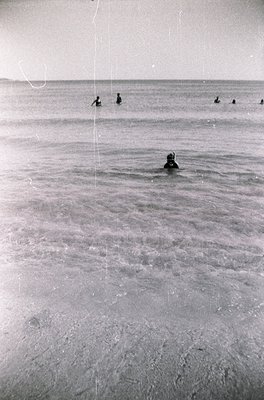 Black-and-white shot of shallow coastal waters with six figures swimming. Foreground swimmer wears a cap; background figures ...