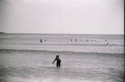 Mid-20th century black-and-white seaside scene: child wading in shallow waves, surrounded by adults swimming in deeper water....