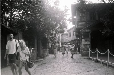 Vintage black-and-white street scene featuring cobblestone pathway flanked by wooden buildings with balconies. Mid-20th centu...