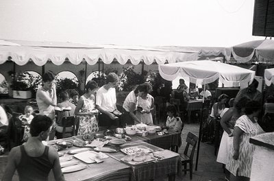 Mid-20th century outdoor café scene with white canopies and floral-patterned dresses. Tables laden with plates, cutlery, and ...