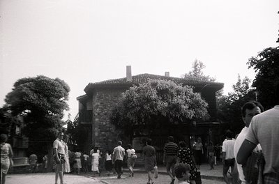 Mid-20th century courtyard scene with stone building featuring rounded corners and arched windows. Crowd of casually dressed ...