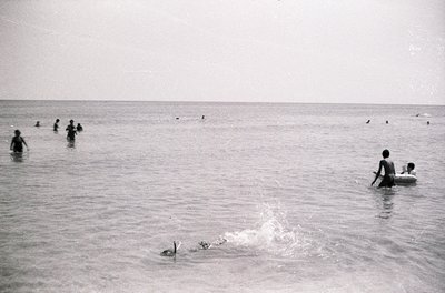 Black-and-white seaside scene with shallow waters, likely mid-20th century. Group of people wading and swimming in calm, open...