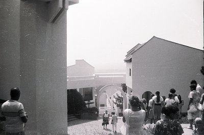Vintage black-and-white courtyard scene with mid-century Brutalist concrete pillar framing left. Group of people in casual 19...