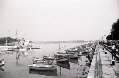 Black-and-white seaside scene featuring a row of small wooden fishing boats docked along a concrete pier. The boats display n...