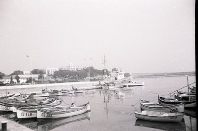 Black-and-white coastal scene featuring a marina with small wooden fishing boats marked with Cyrillic letters/numbers. Larger...