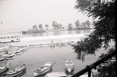 Black-and-white harbor scene featuring small wooden boats docked along a calm waterfront. A mid-20th century passenger ferry ...