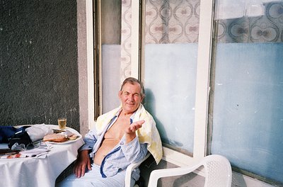 Mid-century man poses indoors with a relaxed gesture, seated in a white plastic chair. A table beside him holds a half-eaten ...