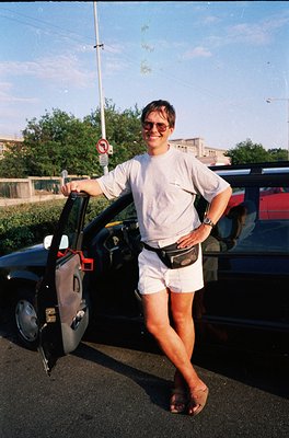 Casual 1990s street portrait: individual in white athletic wear (shirt, shorts, flip-flops) poses by a vintage hatchback car,...