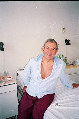 Portrait of an elderly man in a hospital bed, wearing a light blue open jacket and maroon pants. The setting appears to be a ...
