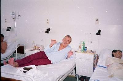 A hospital ward from the 1990s–2000s, featuring three patients in shared beds. Central figure reclines in a light blue hospit...