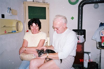 A mid-20th-century indoor scene featuring an elderly couple bonding with a small black-and-tan dog. The man, wearing glasses ...