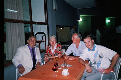 Four men seated at a table in a dimly lit indoor setting, likely a 1980s-1990s restaurant or lounge. The man on the left wear...