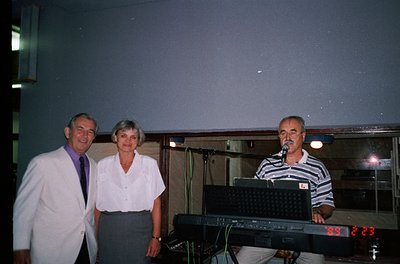 Indoor event featuring three adults posing near a keyboard with digital clock displaying "23:23." Man on left wears a white l...