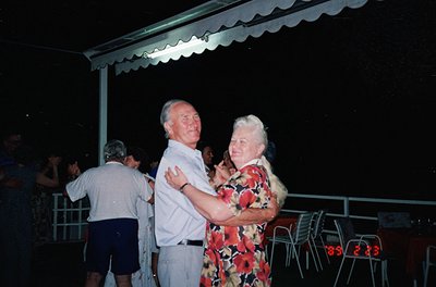 Couple dancing under a wavy-patterned awning at an outdoor night event, likely a 1980s-1990s seaside resort. Man in white shi...