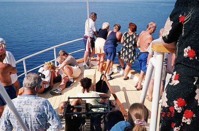 Mid-1970s cruise ship deck scene: adults in retro swimwear (one-man suit, floral dresses) gather on sun deck by railing, some...