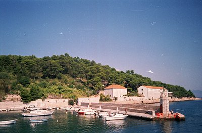 Coastal village scene with stone buildings perched on a hillside, overlooking a small harbor. White boats docked at a stone p...