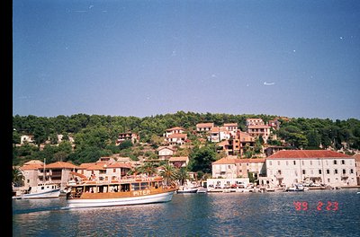 Mediterranean coastal village with clustered stone buildings and terracotta roofs. A vintage ferry labeled "Piter" docked nea...