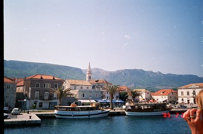 Mediterranean coastal town with stone buildings, palm trees, and a church steeple. Two boats docked near the waterfront, one ...