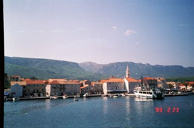 Mediterranean coastal village with stone buildings, terracotta roofs, and a prominent church steeple. Small boats docked alon...