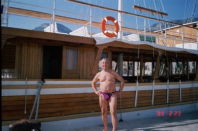 Vintage seaside scene featuring a shirtless man in 1970s-style swim trunks posing beside a wooden docked boat. The vessel dis...