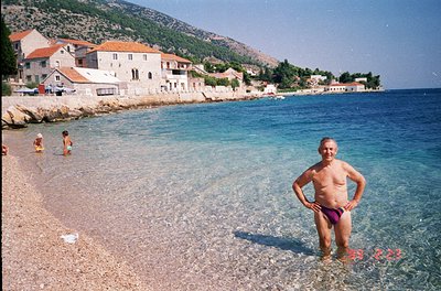 Vintage seaside scene featuring a man in red swim trunks standing in shallow turquoise waters, with Mediterranean-style stone...