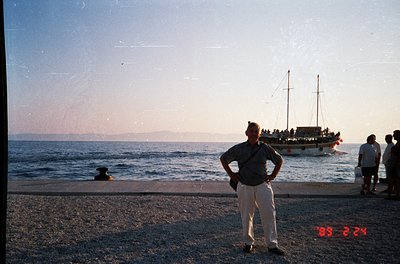 Vintage seaside scene with man in light-colored trousers and dark polo standing on pebble shore. Wooden pier extends into cal...