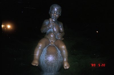 Bronze statue of a seated child playing a flute atop a spherical base, likely a public art piece. The low-angle lighting enha...