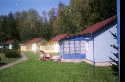 Vintage row of pastel-colored beach huts with red roofs, lined along a grassy path. Bright blue doors and windows contrast wi...
