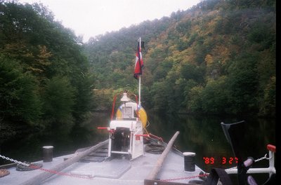 Vintage ferry crossing a narrow, forested river with autumn foliage. White vessel marked with red life rings and a red/white ...