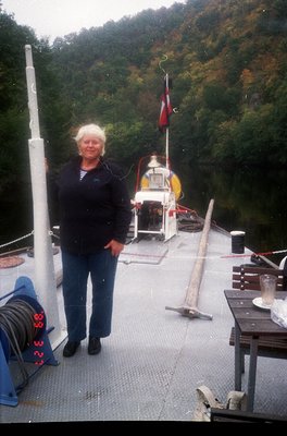 Woman in casual 1990s attire stands on a small wooden ferry deck, near a red-and-white flag. The vessel features a simple cab...