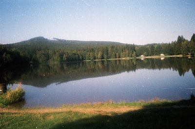 Serene alpine lake nestled between forested hills, reflecting clear skies and distant structures. Calm waters mirror surround...