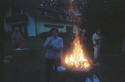 Vintage evening gathering around a bonfire in a residential backyard, likely 1970s–1980s. Group of adults in casual attire (t...