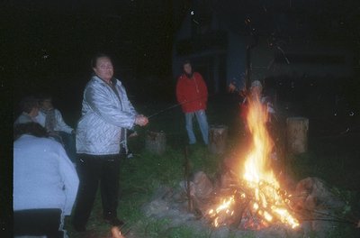 Nighttime outdoor gathering around a roaring bonfire. Four individuals in casual winter attire (puffer jackets, jeans) roast ...