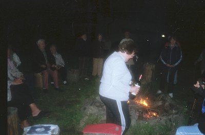 Nighttime outdoor gathering around a small bonfire. A person in a white jacket and black pants lights a candle or small firew...