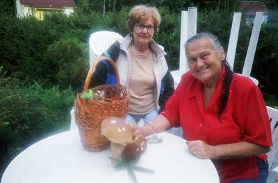 Two individuals seated outdoors at a white table, holding a basket of foraged mushrooms. The woman on the left wears glasses ...