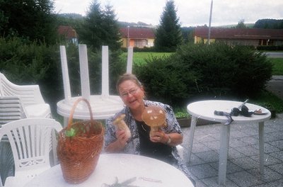 A woman poses outdoors at a café-style table holding two pastries. White plastic chairs and tables surround her, with a woven...