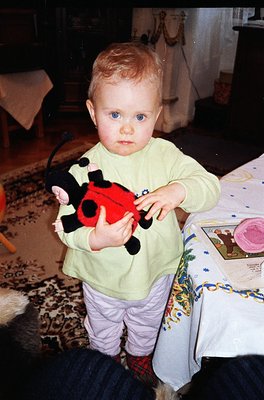 Vintage-style photo of a toddler (approx. 1-2 years) holding a plush Ladybug toy in a cozy indoor setting. Patterned rug, flo...