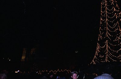 Vintage nighttime festive scene with illuminated tree and blurred crowd. Warm string lights outline a tall, conical structure...