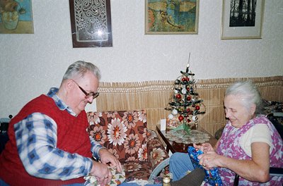 Two elderly individuals crafting in a cozy 1970s-era living room. The man, wearing a plaid sweater and glasses, and the woman...