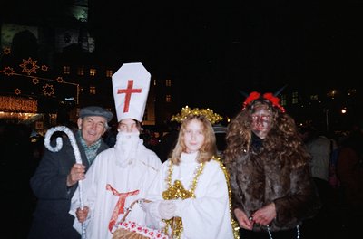 Four individuals in festive holiday costumes pose under illuminated city lights. From left: a man in a faux-fur hat and coat ...