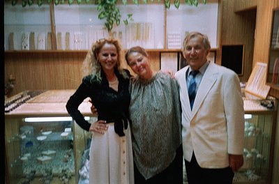 Three individuals pose indoors in a well-lit, mid-century setting. The woman on the left wears a sleeveless black top with a ...