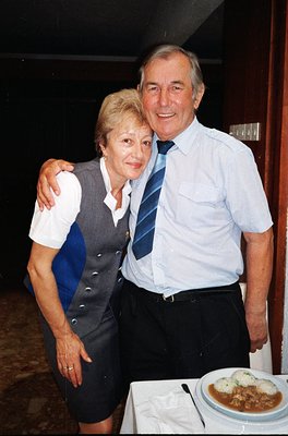 Couple posing indoors at a formal event, likely 1990s–2000s. Woman in a gray vest over a white top, man in a light blue dress...