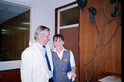 Two individuals in a formal indoor setting, likely a 1980s-1990s office or hotel lobby. The man wears a white lab coat and st...