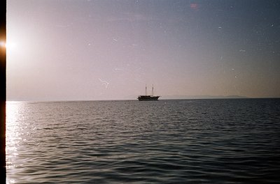 Vintage seascape shot of a single-masted wooden sailing vessel on calm waters, framed by a window sill. Soft, grainy film gra...