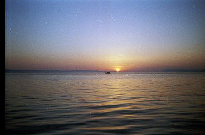 Sunset over calm waters with a lone boat silhouetted near horizon. Warm golden light reflects on rippling sea, framed by fadi...