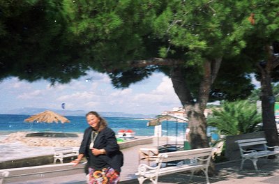 Vintage seaside scene featuring a woman in a patterned dress walking under a large tree, with ocean and beach umbrellas in ba...